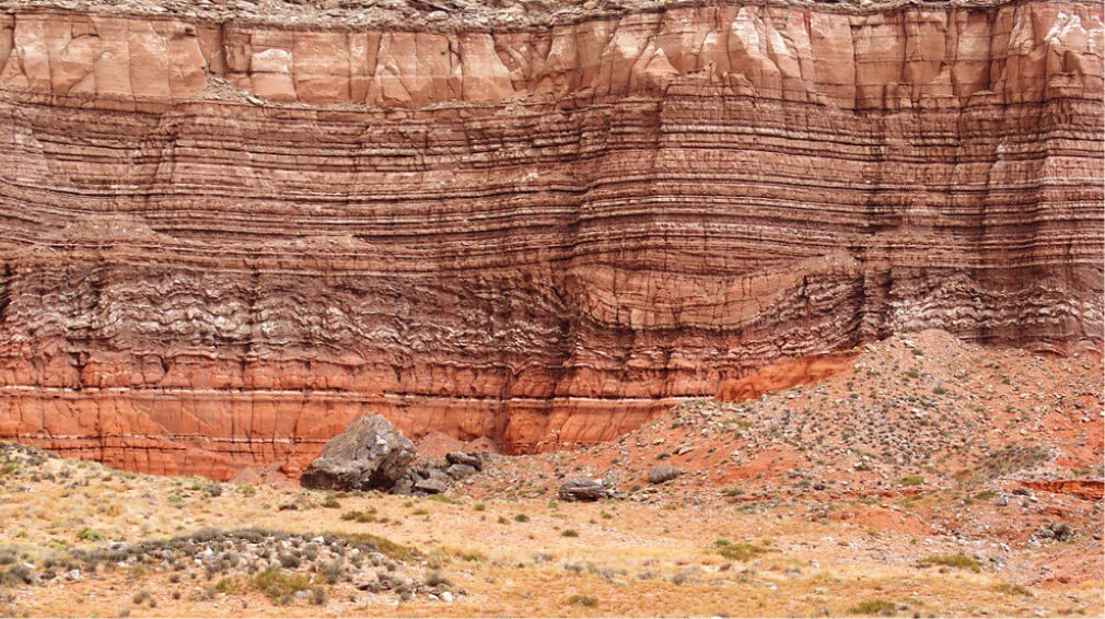 Layered rock formations in a desert landscape with sparse vegetation and scattered rocks.