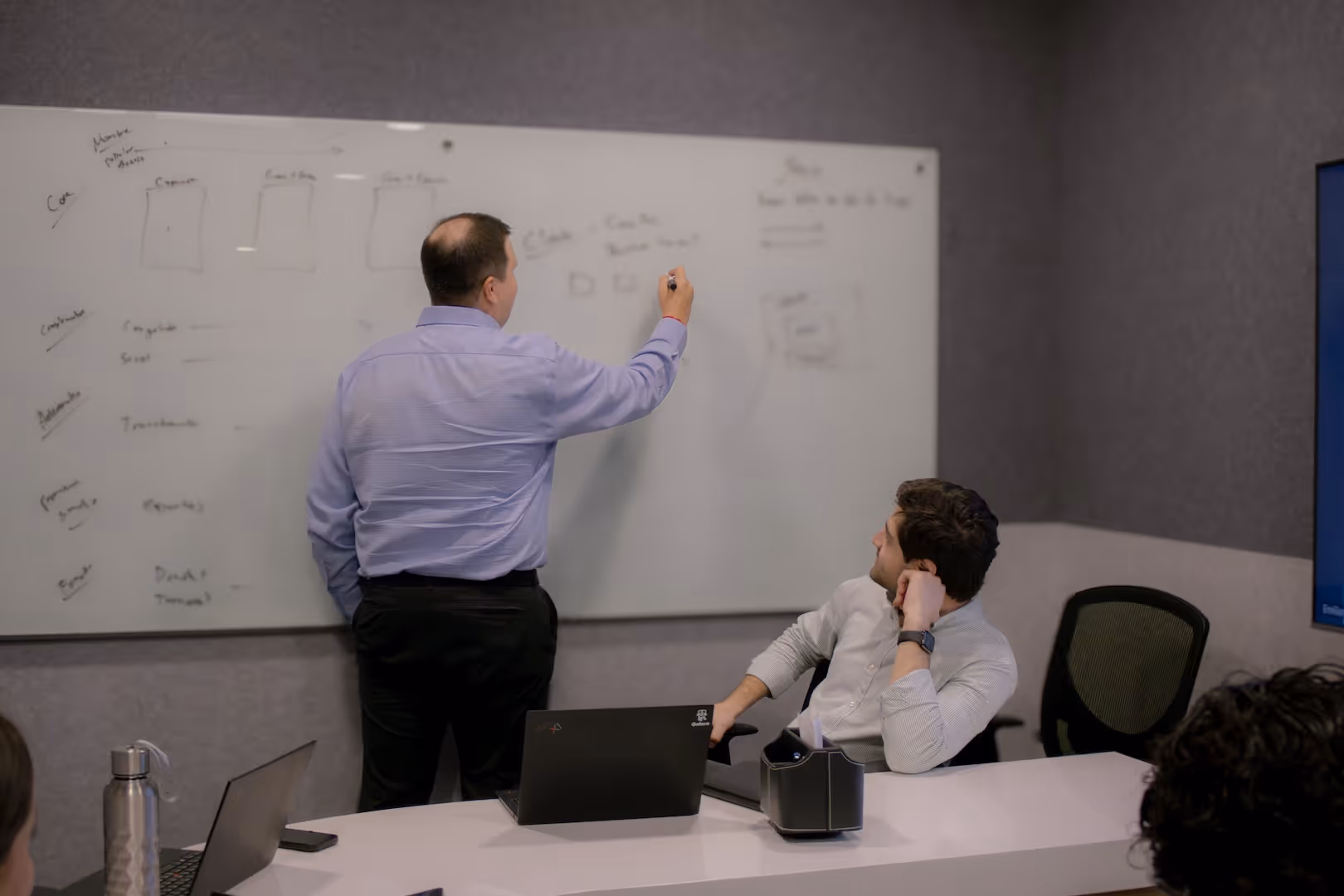 A man in a light blue shirt writing on a whiteboard while a seated man attentively watches in a meeting room.
