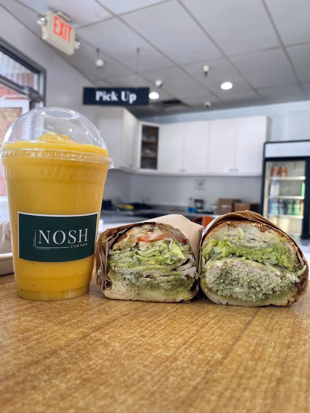 Two halves of a sandwich with lettuce and creamy green spread next to a yellow smoothie in a clear cup labeled NOSH CORNER on a wooden table inside a cafe.