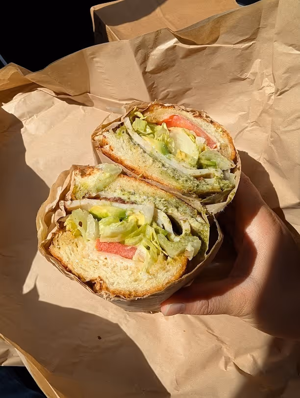 Hand holding a sandwich cut in half with lettuce, tomato, cheese, and green pesto sauce on a crusty white bread wrapped in brown paper.