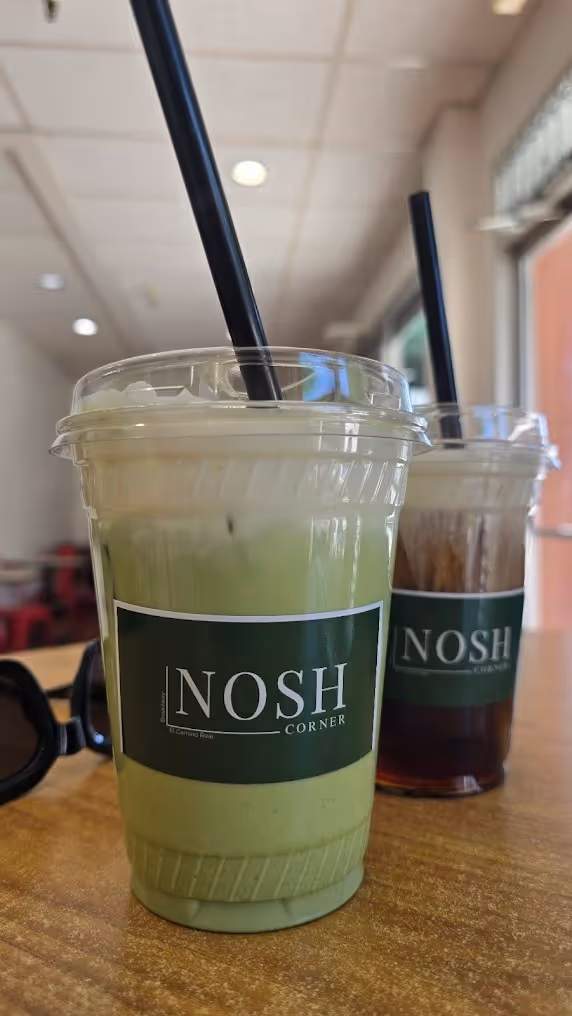 Two cold beverages in plastic cups with black straws on a table, one green matcha drink in front and a darker drink behind, both labeled 'NOSH Corner'.