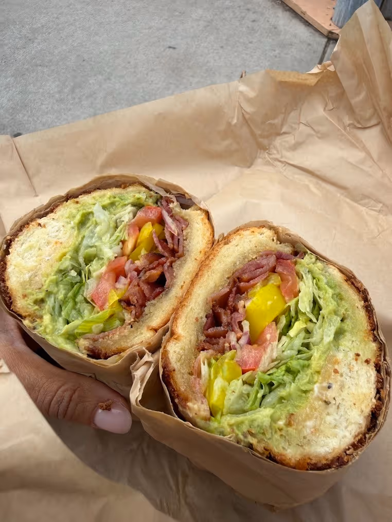 Close-up of a hand holding a sandwich cut in half with bacon, lettuce, tomato, and yellow peppers wrapped in brown paper.