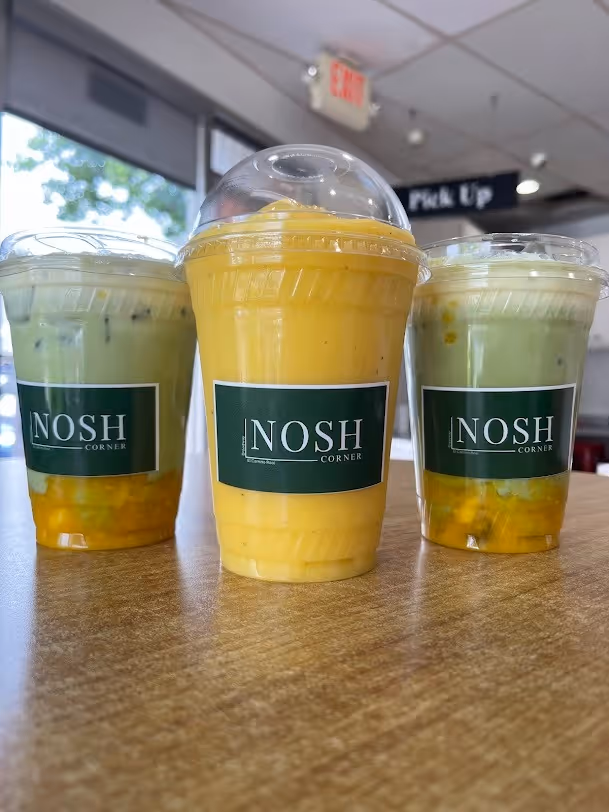 Three plastic cups with clear lids filled with yellow and green drinks labeled 'NOSH Corner' on a wooden table in a cafe setting.