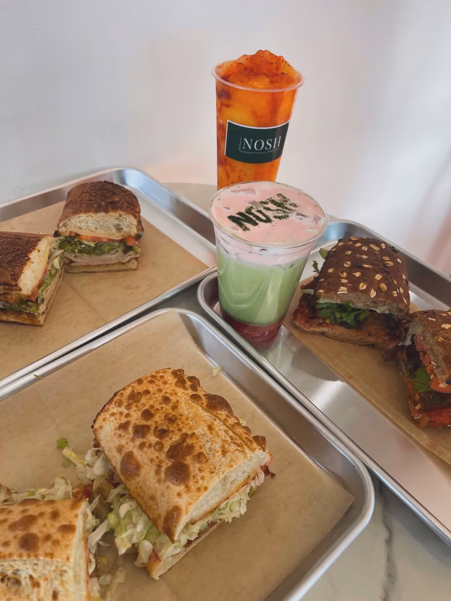 Three toasted sandwiches with various fillings on trays and two colorful drinks, one labeled NOSH and another with pink foam topped with green powder.