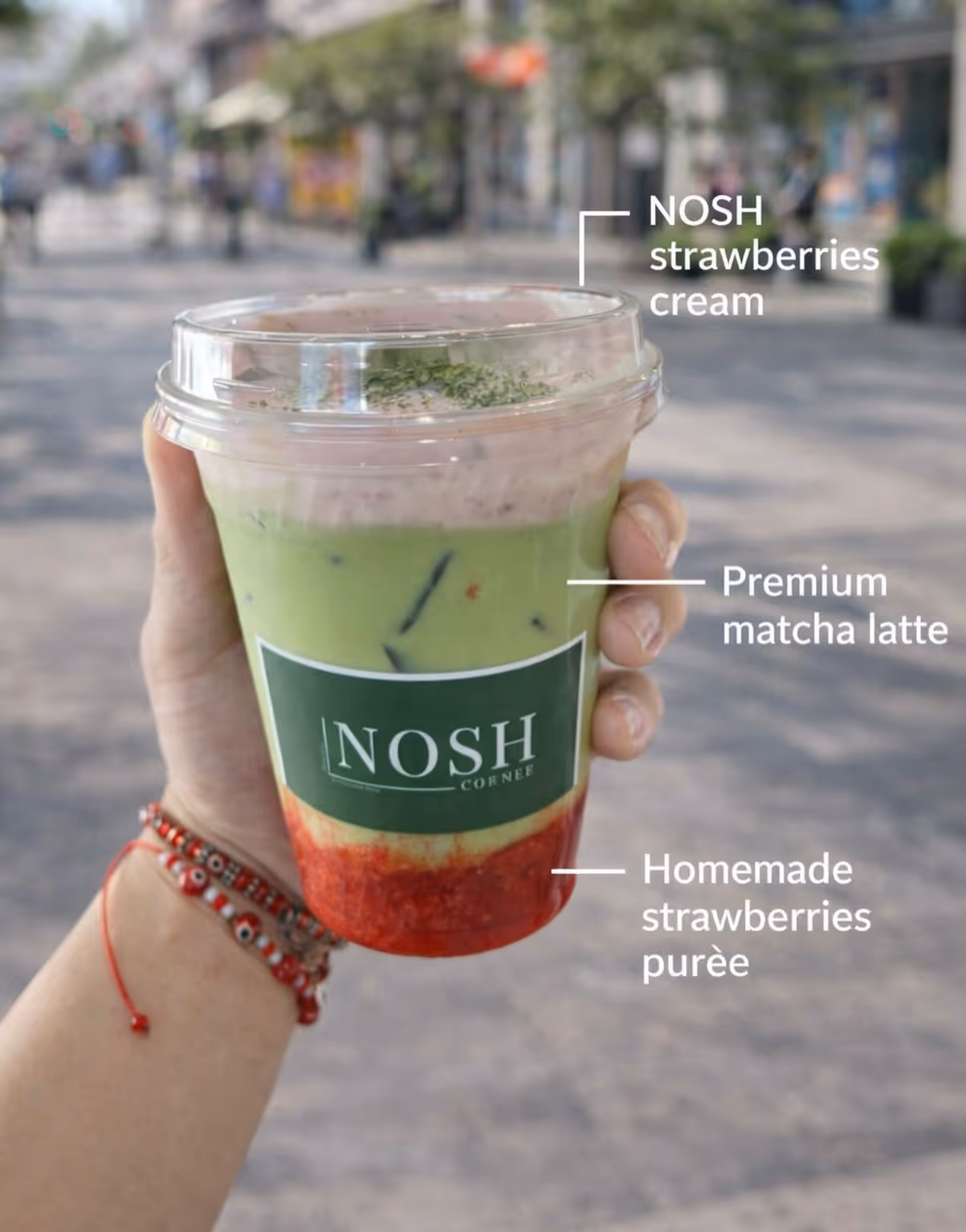 Hand holding a NOSH Cornee branded cup with layered drink of homemade strawberries purée, premium matcha latte, and NOSH strawberries cream on top.