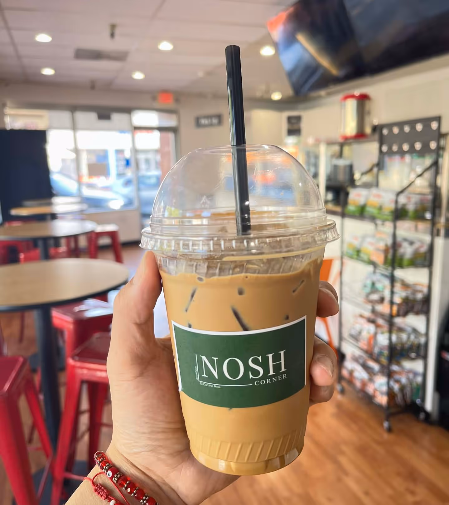 Hand holding an iced coffee in a plastic cup with a dome lid and black straw inside a cafe with red stools and a snack rack.