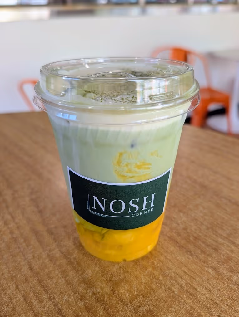 Plastic cup with clear lid containing a layered green and yellow drink with a black NOSH Corner label on the front, placed on a wooden table.