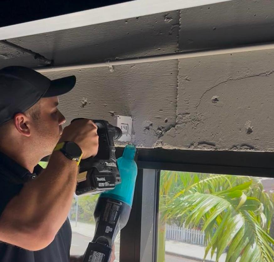 Technician drilling into a concrete ceiling above a window to mount a shade bracket