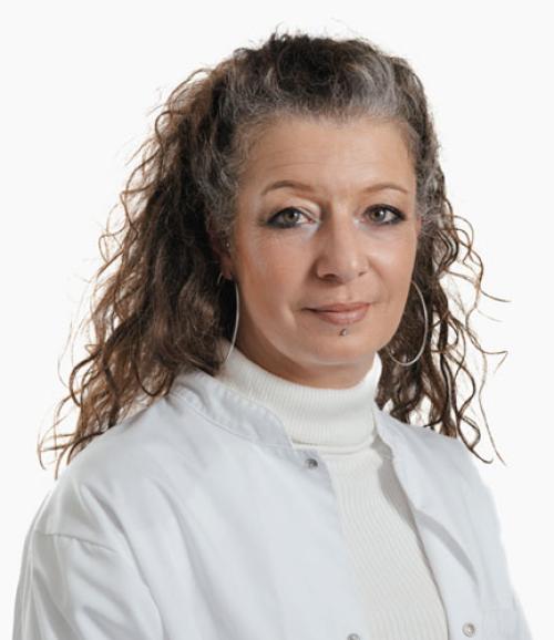 Porträt einer Frau mit lockigem Haar, die einen weissen Rollkragenpullover und eine weisse Jacke trägt, mit grossen silbernen Creolen als Schmuck.