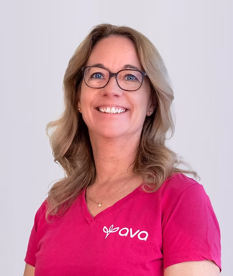 Fröhliche Frau mit blonden, leicht gewellten Haaren, die eine rote Ava-T-Shirt und eine Brille trägt, vor hellem Hintergrund.