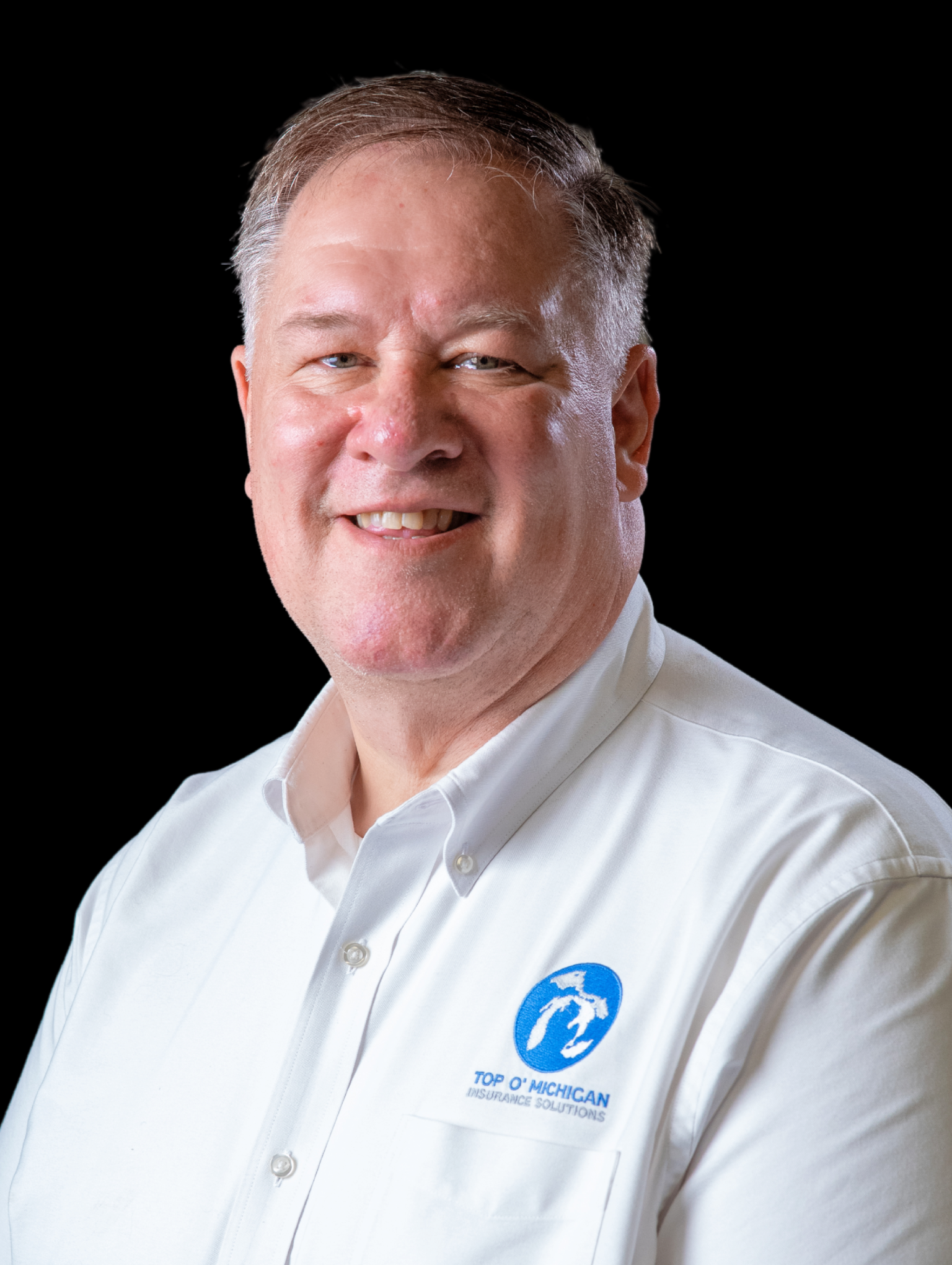 Smiling middle-aged man in a white shirt with Top O' Michigan Insurance Solutions logo on the chest against a black background.
