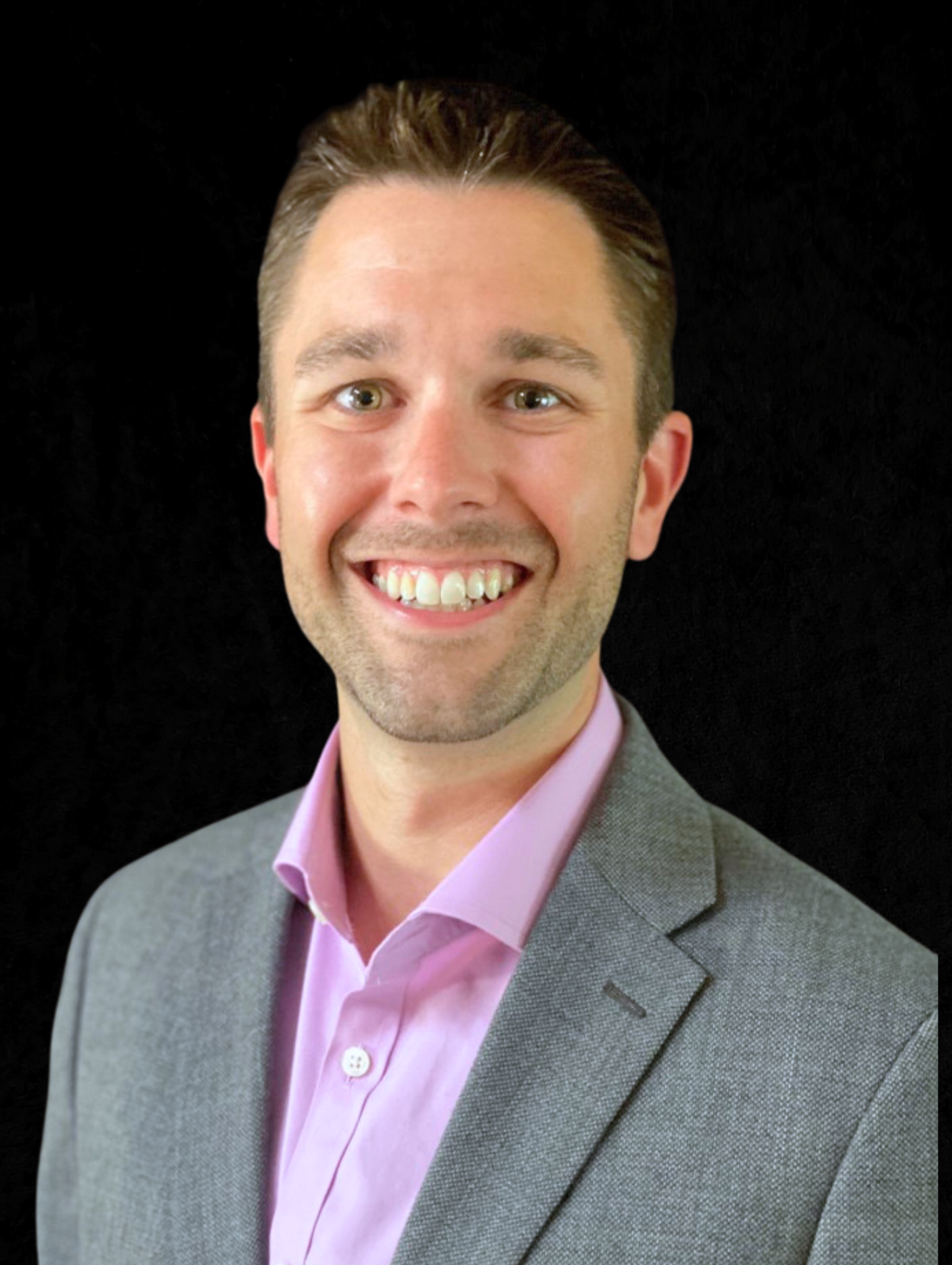 Smiling man with short brown hair wearing a gray blazer and light purple dress shirt against black background.