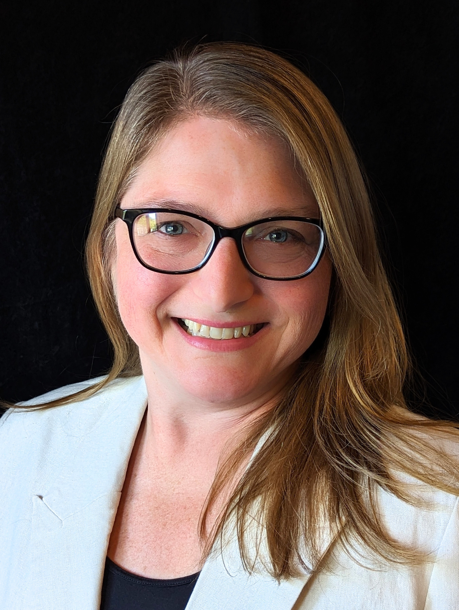 Smiling woman with glasses, long light brown hair, wearing a white blazer and black top, against a black background.