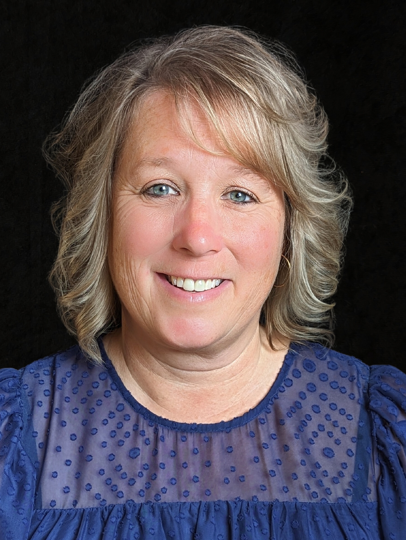 Smiling woman with curly blonde hair wearing a blue textured blouse against a black background.