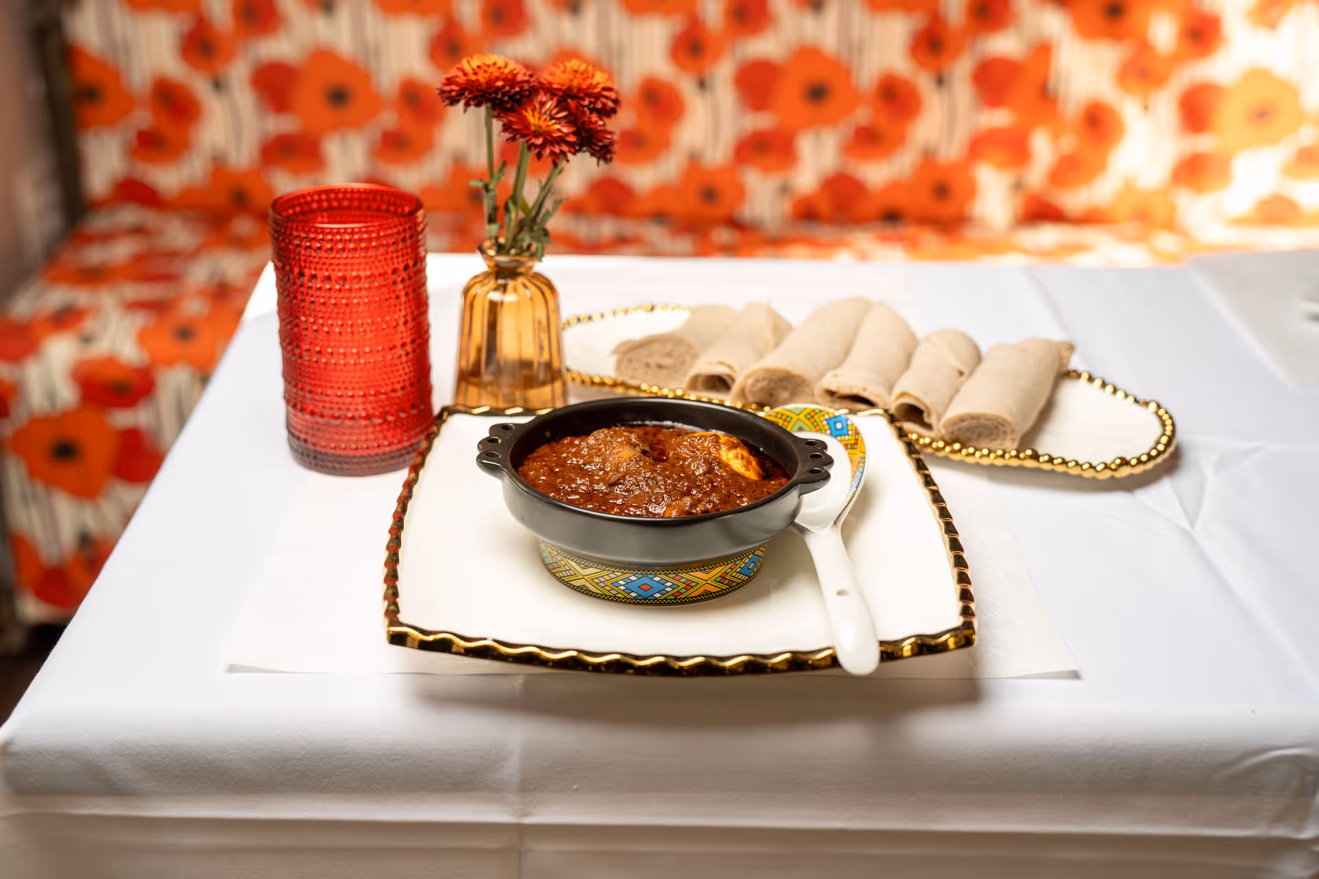 Ethiopian injera bread with a bowl of spicy stew on a white tablecloth with a red glass and small vase with red flowers.