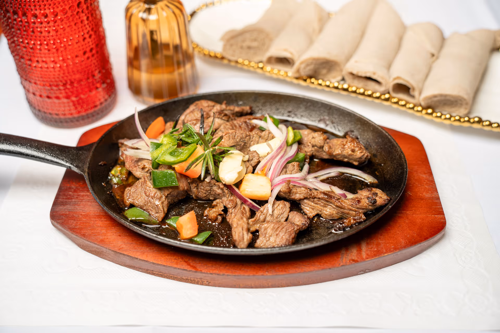 Cast iron skillet with cooked beef strips, bell peppers, onions, and herbs, served on a wooden board with rolled bread and drinks in the background.