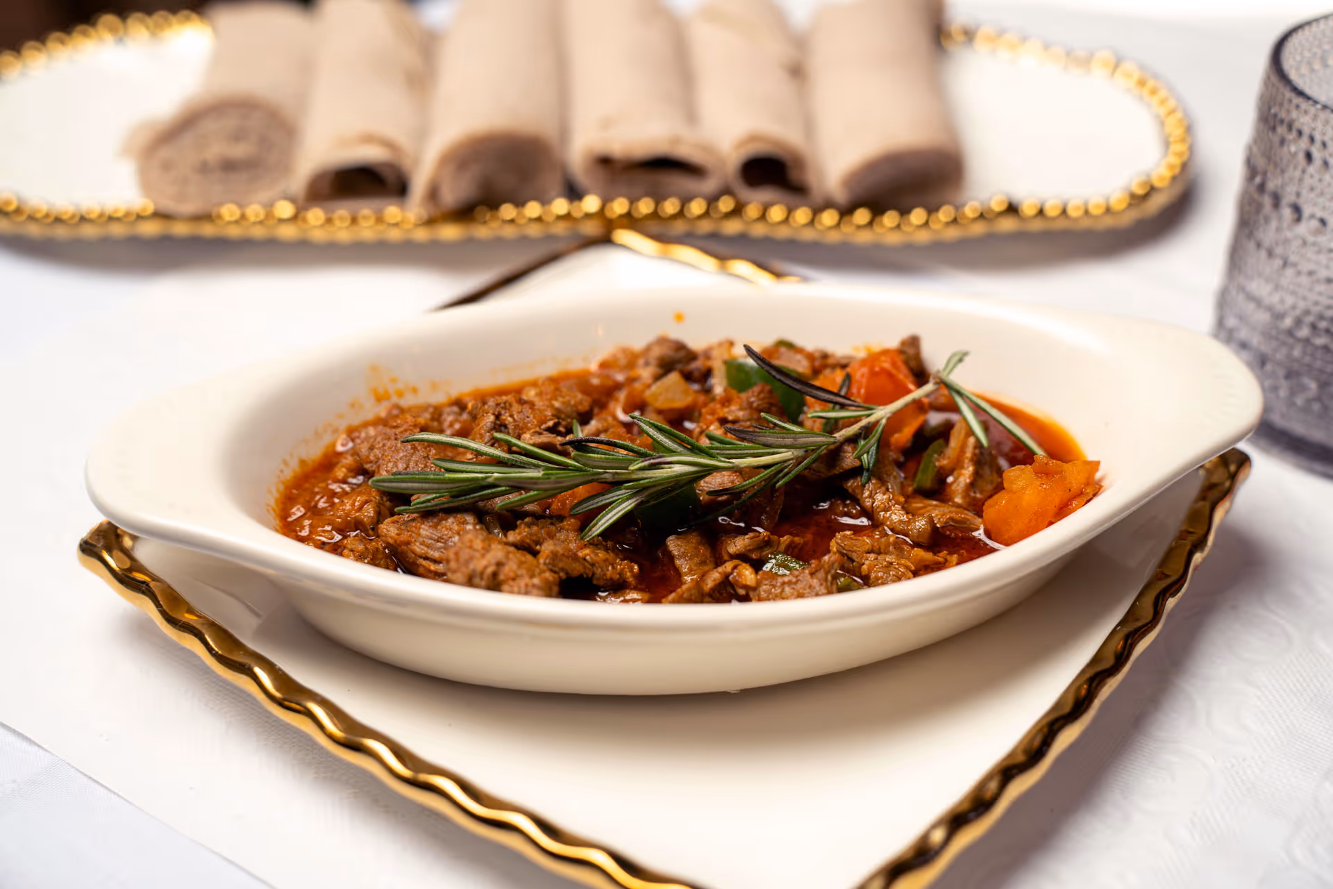 Bowl of beef stew garnished with rosemary, served with rolled bread on a decorative tray nearby.