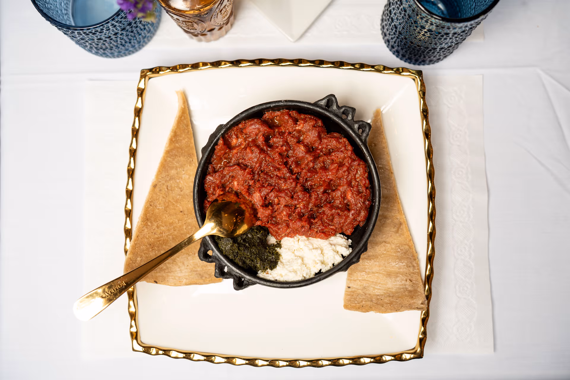 Traditional Ethiopian kitfo with red spiced minced meat, white cheese, and green herb sauce served with pieces of injera bread on a white and gold-rimmed plate.