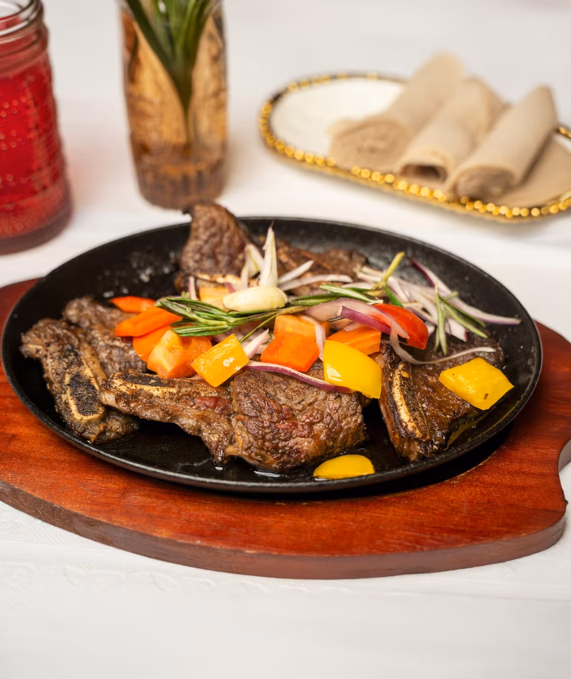 Grilled beef ribs garnished with chopped carrots, yellow bell peppers, red onions, and rosemary on a black skillet over a wooden platter.