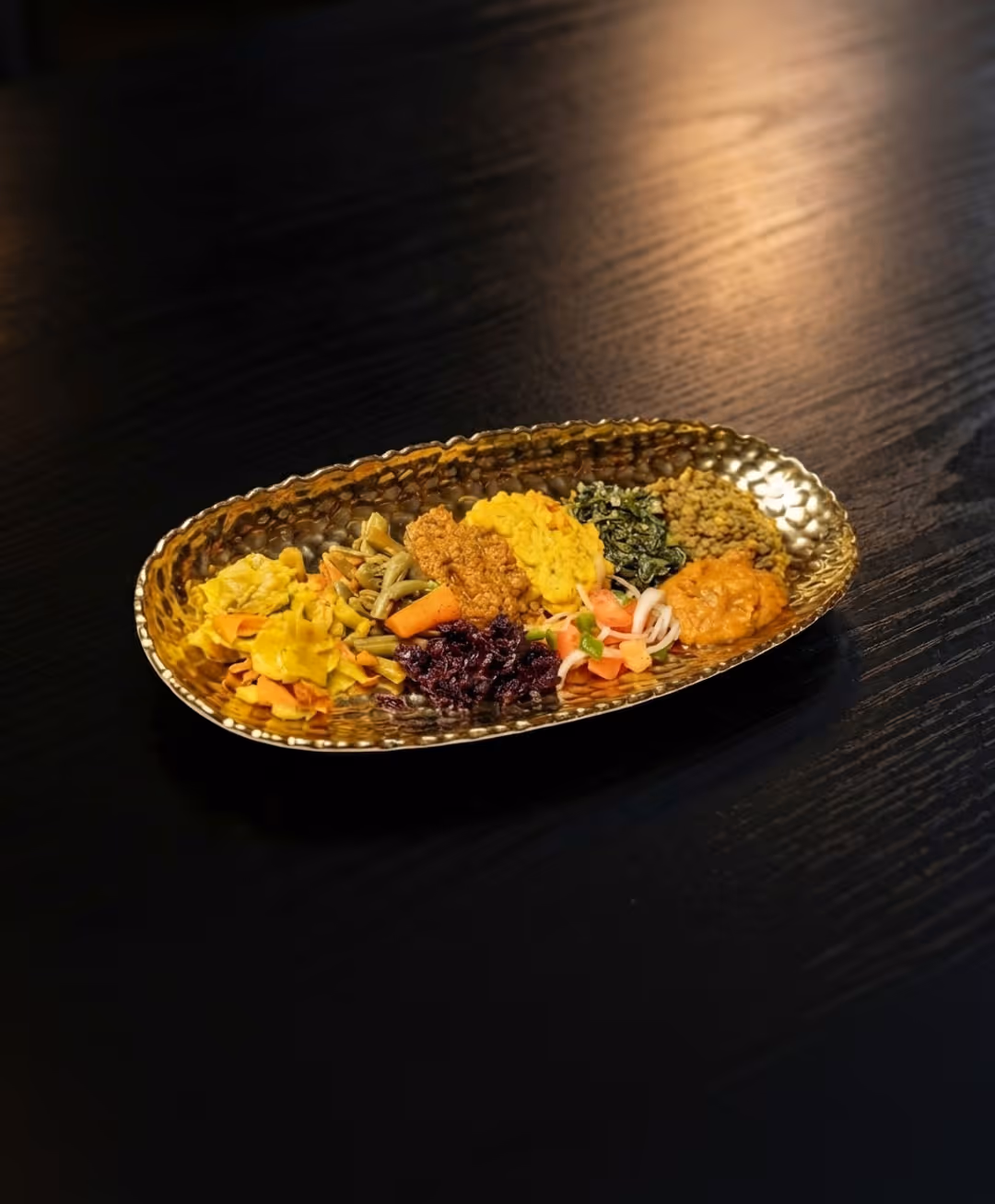 Assorted colorful vegetable and lentil dishes arranged on an oval hammered brass plate on a dark wooden table.