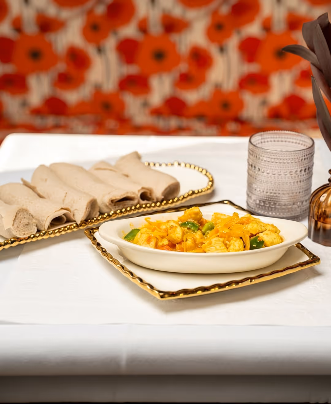 Table setting with a bowl of vegetable stew, rolled injera on a tray, a textured tumbler, and a vase with dark leaves against a red floral background.
