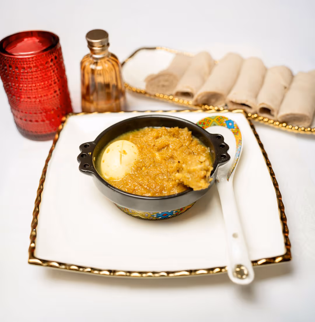 Bowl of Ethiopian doro wat stew with a boiled egg on a decorative plate, accompanied by rolled injera bread, a red candle, and a perfume bottle.