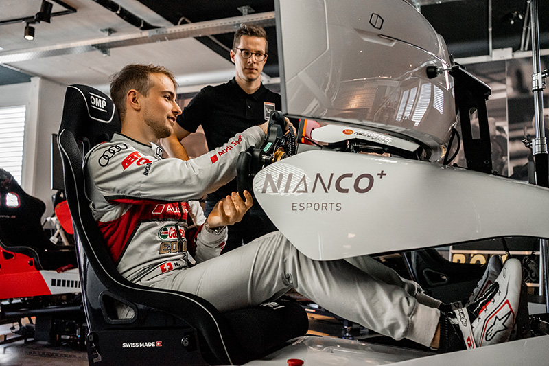 Mann in Rennanzug sitzt in NIANCO+ Esports-Rennsimulator, während ein anderer Mann zuschaut.
