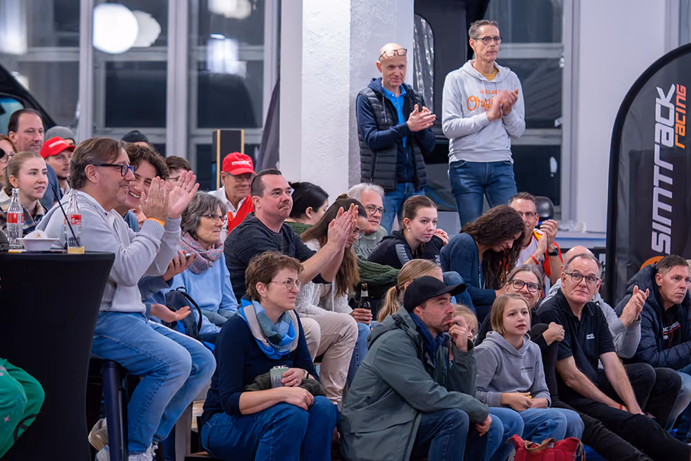 Eine Gruppe von Menschen sitzt und steht in einem Innenraum bei einer Veranstaltung, einige klatschen, andere beobachten aufmerksam, eine Fahne mit der Aufschrift 'Simtrack Racing' ist sichtbar.