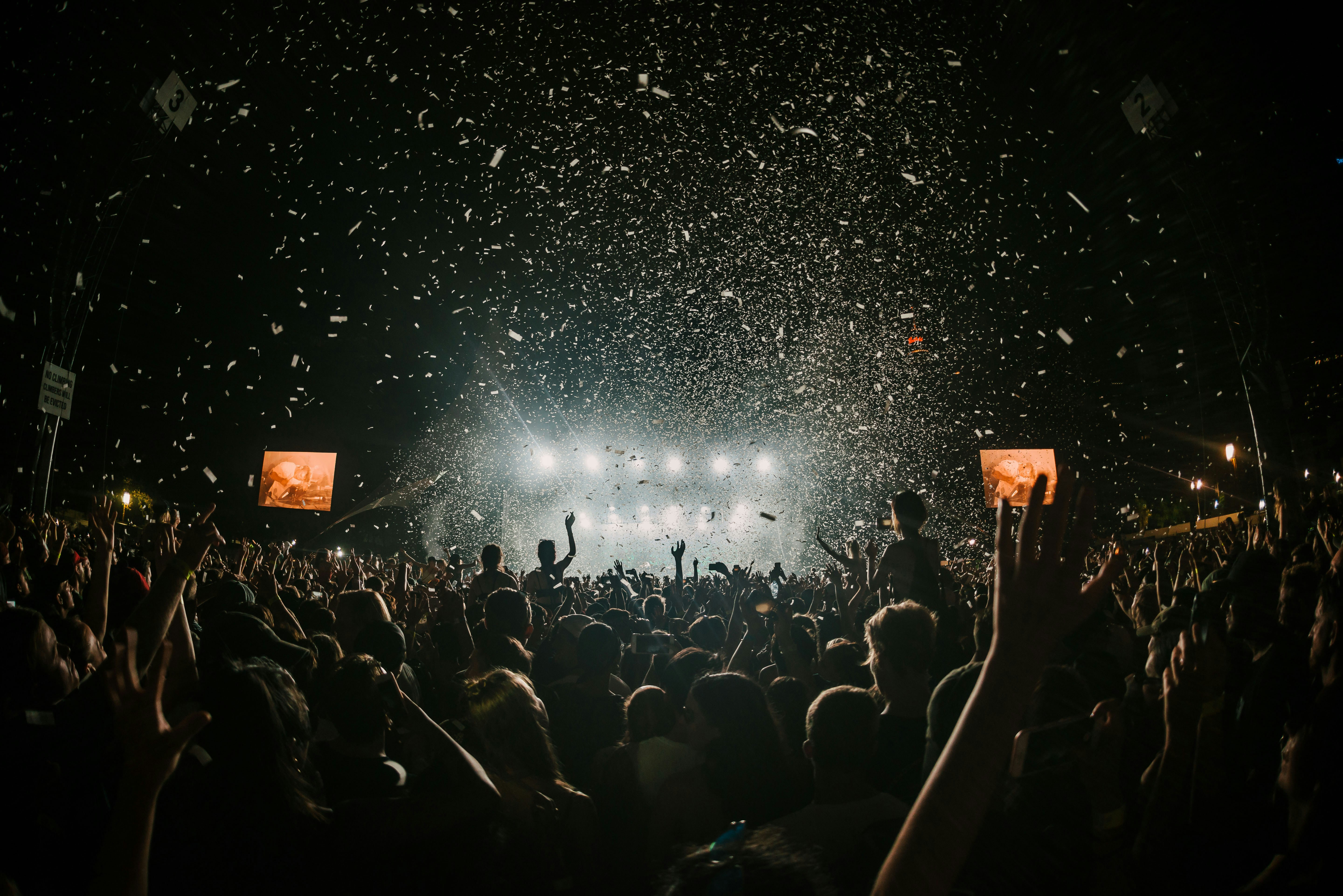 Large crowd at a nighttime concert with confetti falling and bright stage lights in the background.