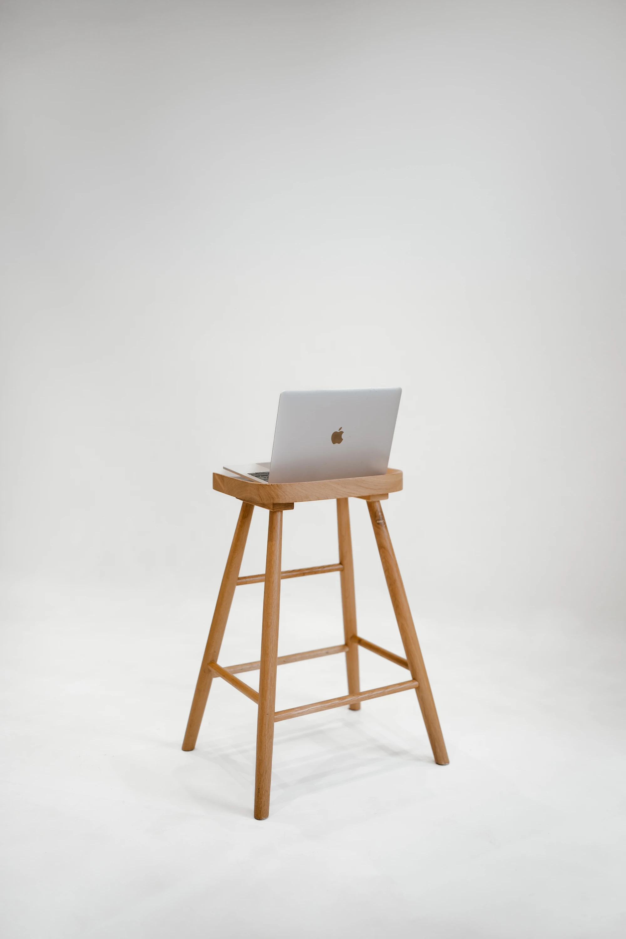 A sleek, silver laptop sits open on a tall, wooden minimalist stool against a plain white background, conveying simplicity and modernity.