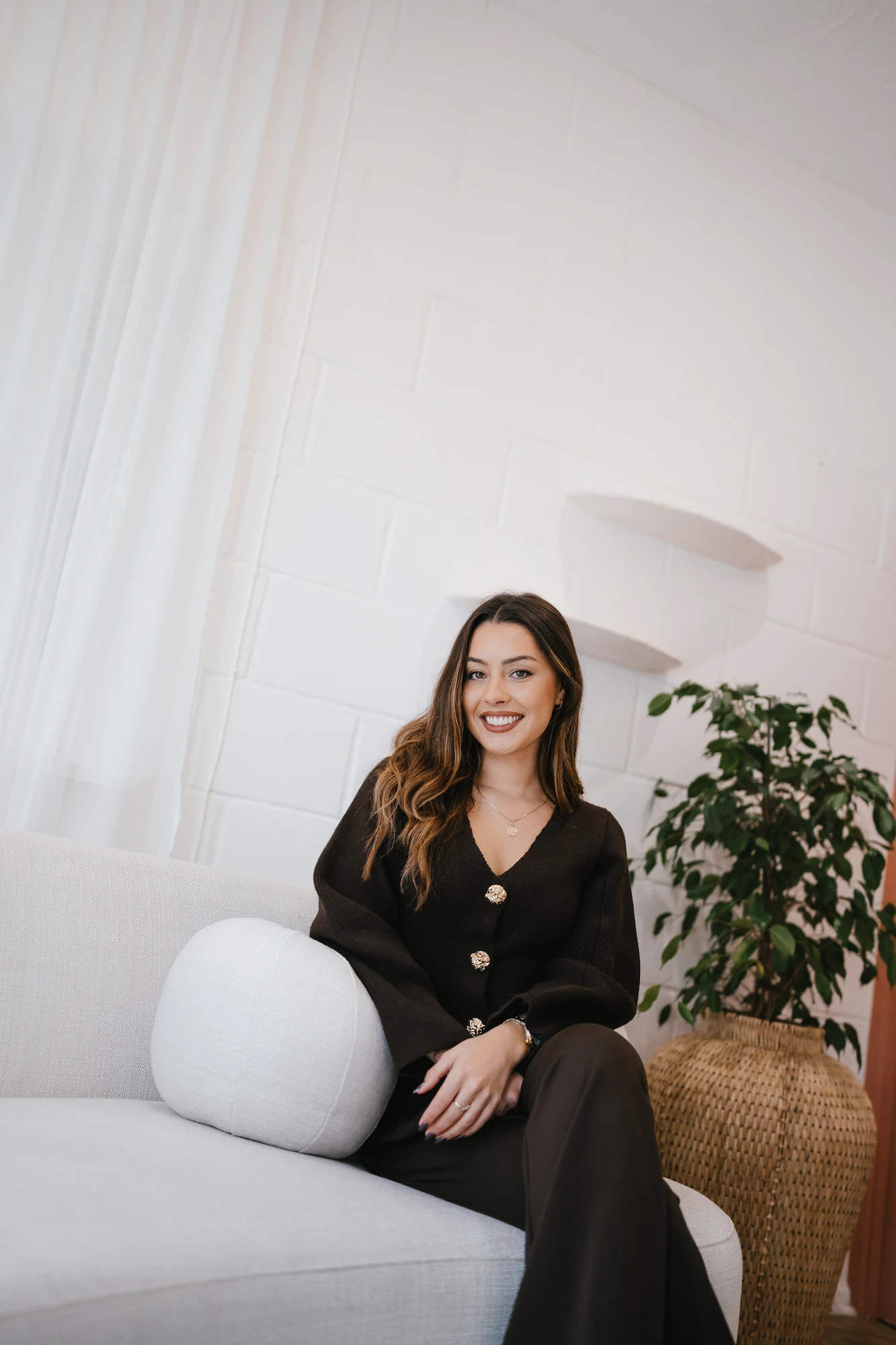 Erin Bayliss, founder of Rinni, in a dark outfit sits on a light sofa in a modern, minimalist room with white brick walls and a potted plant, conveying warmth and elegance.