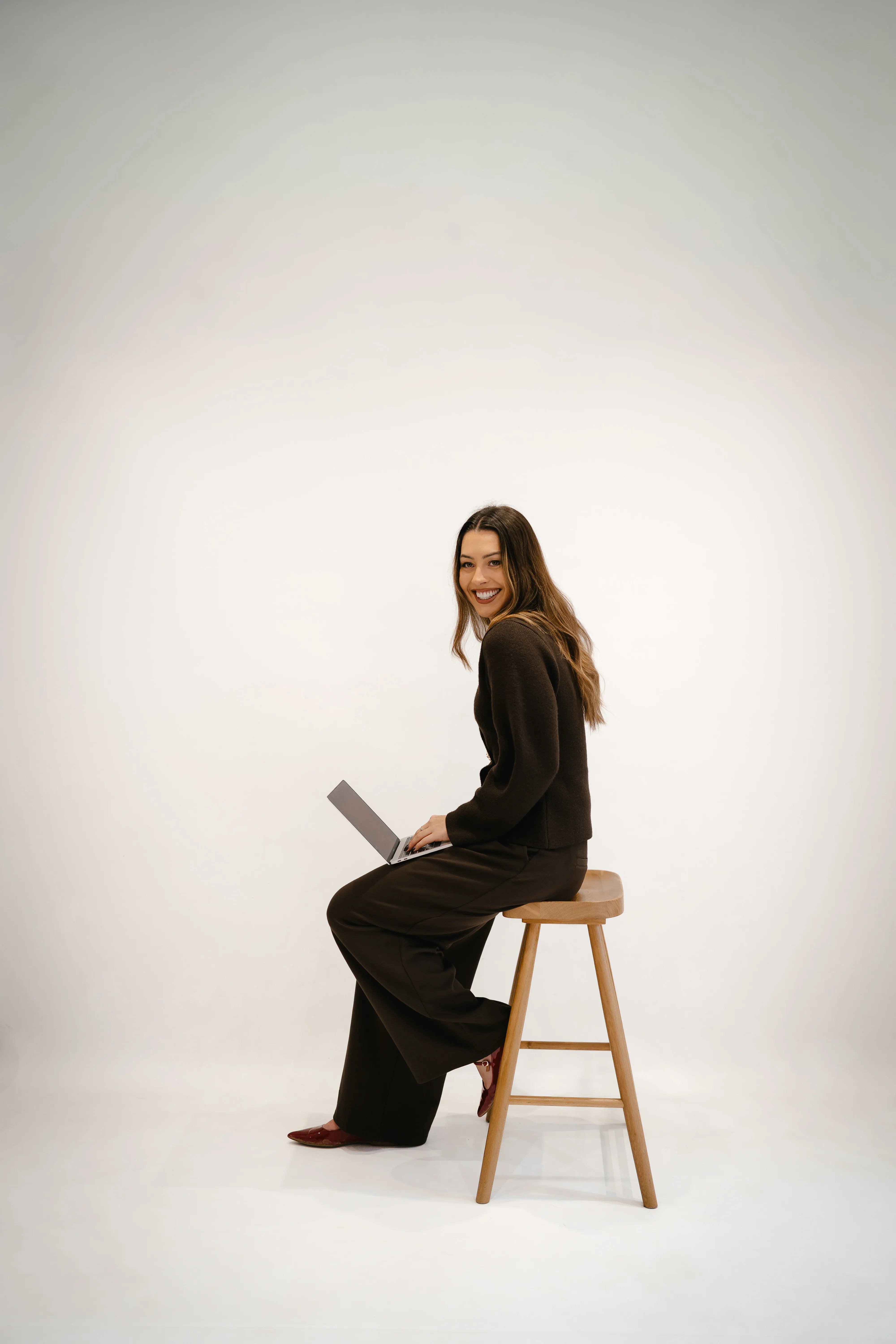 Erin Bayliss, founder of Rinni, sits on a wooden stool against a white background, smiling warmly while holding a laptop. She is wearing a brown outfit and red shoes.