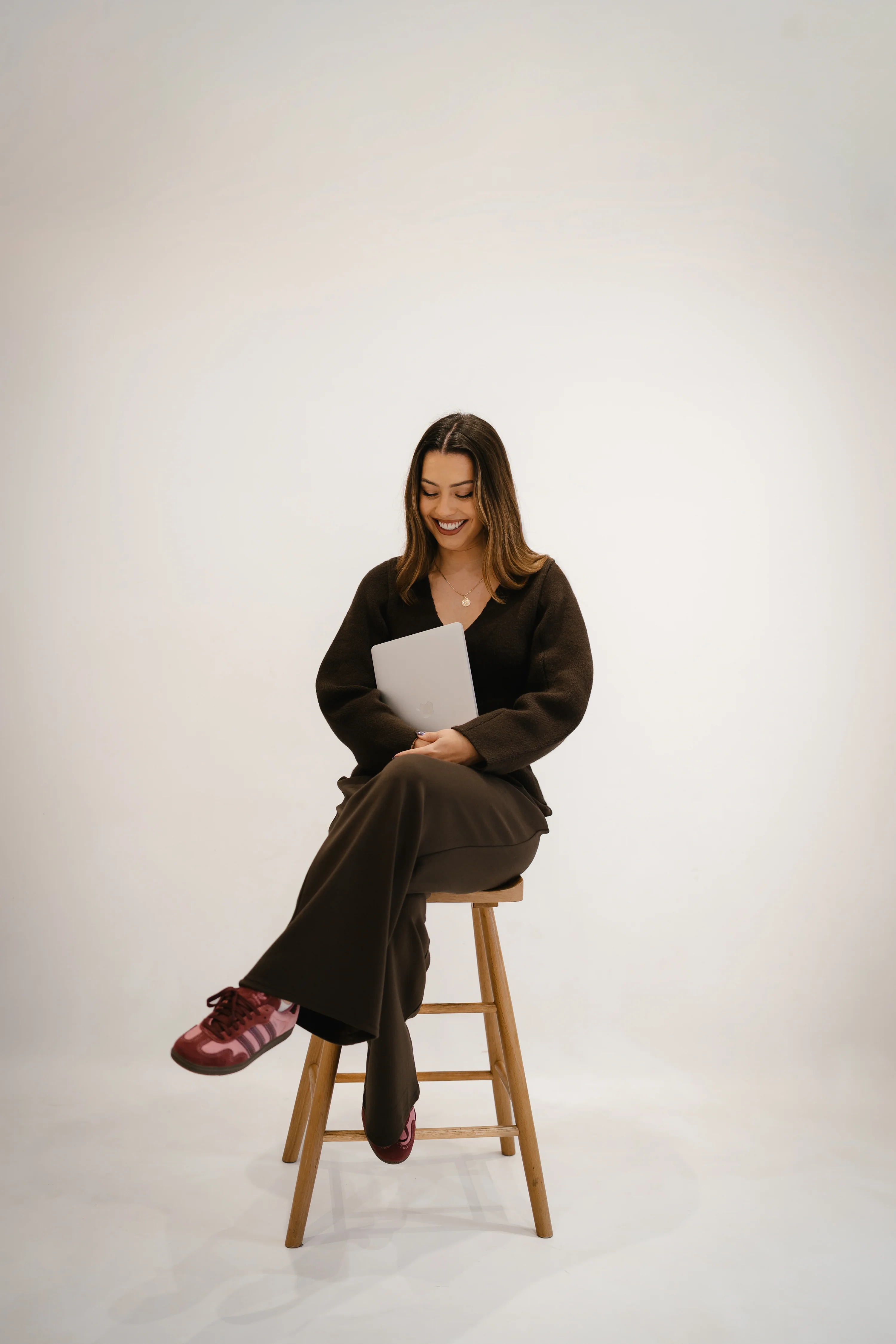 Erin Bayliss, founder of Rinni, in a black sweater and trousers sits on a wooden stool, holding a white folder. She wears red sneakers against a plain background.