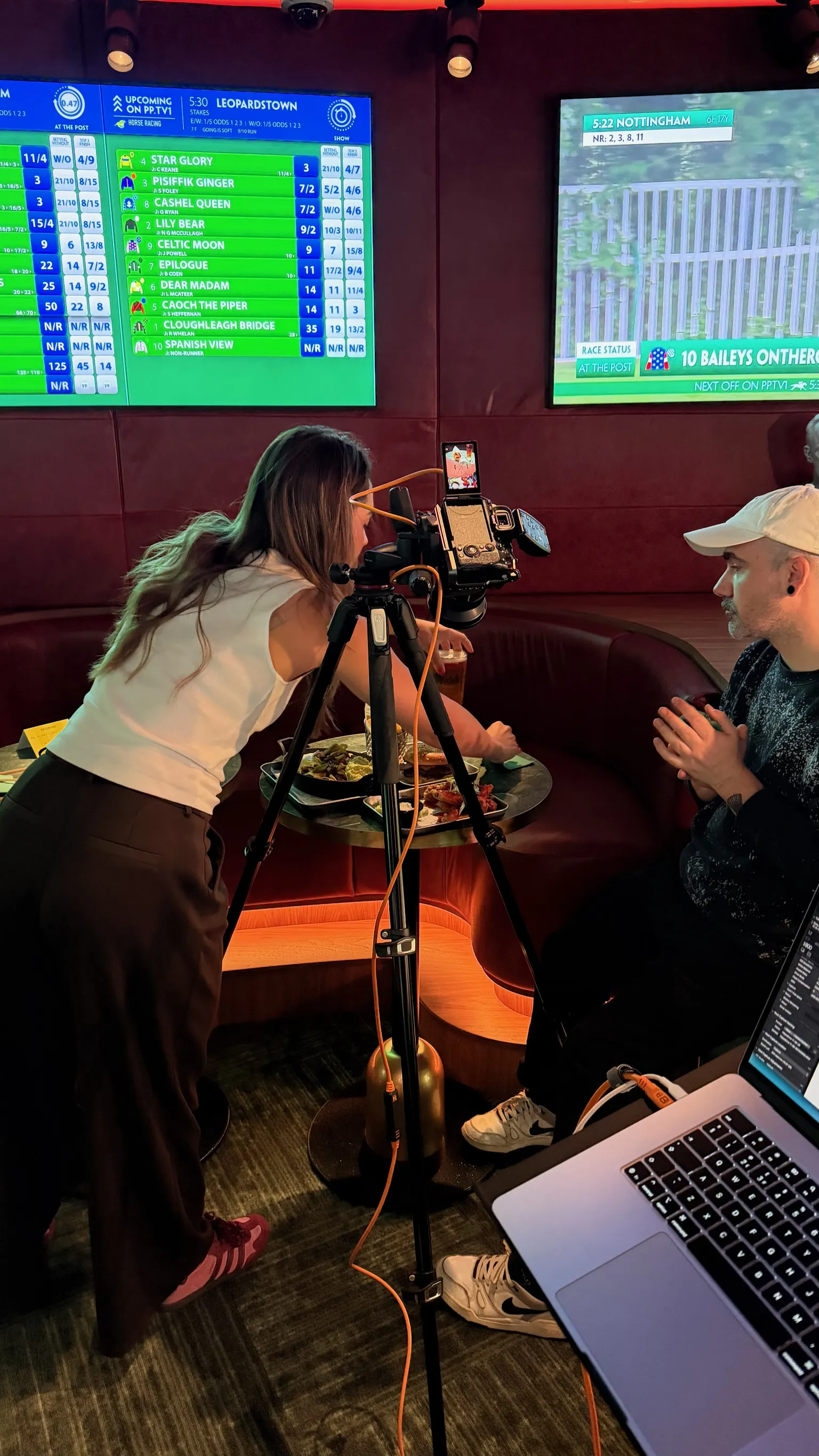 Erin Bayliss adjusting a camera on a tripod aimed at food on a table in a sports lounge. A man watches, seated nearby with TV screens in the background, ready to film.