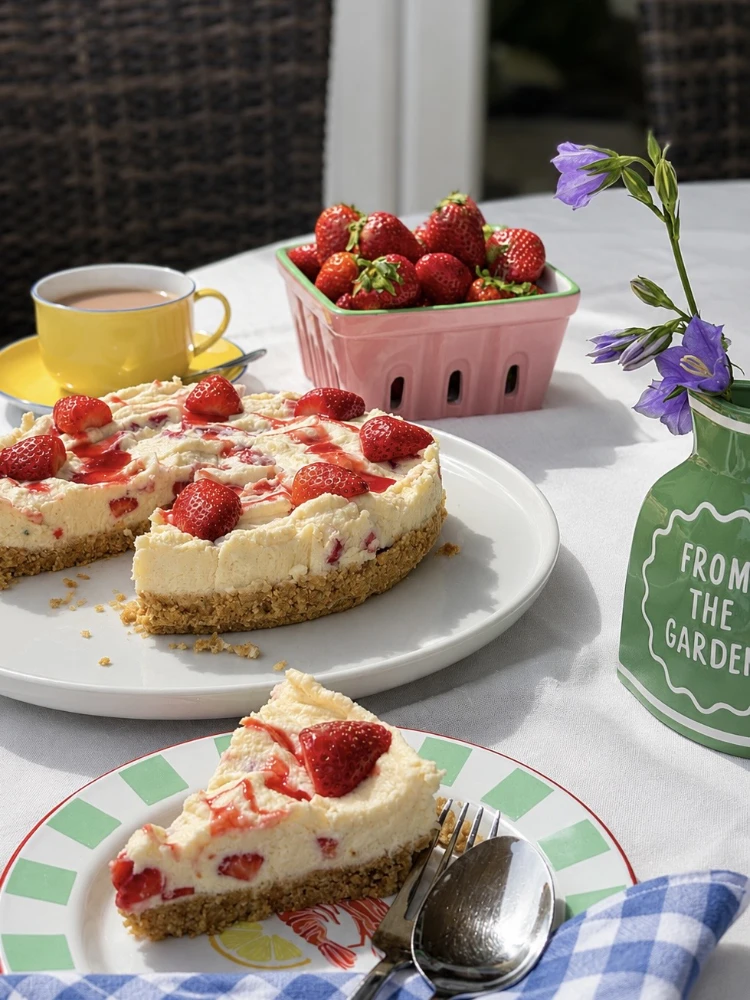A creamy cheesecake adorned with strawberries and syrup sits on a table with a slice on a plate. A basket of strawberries, a yellow cup, and purple flowers in a green vase add a fresh, vibrant touch.