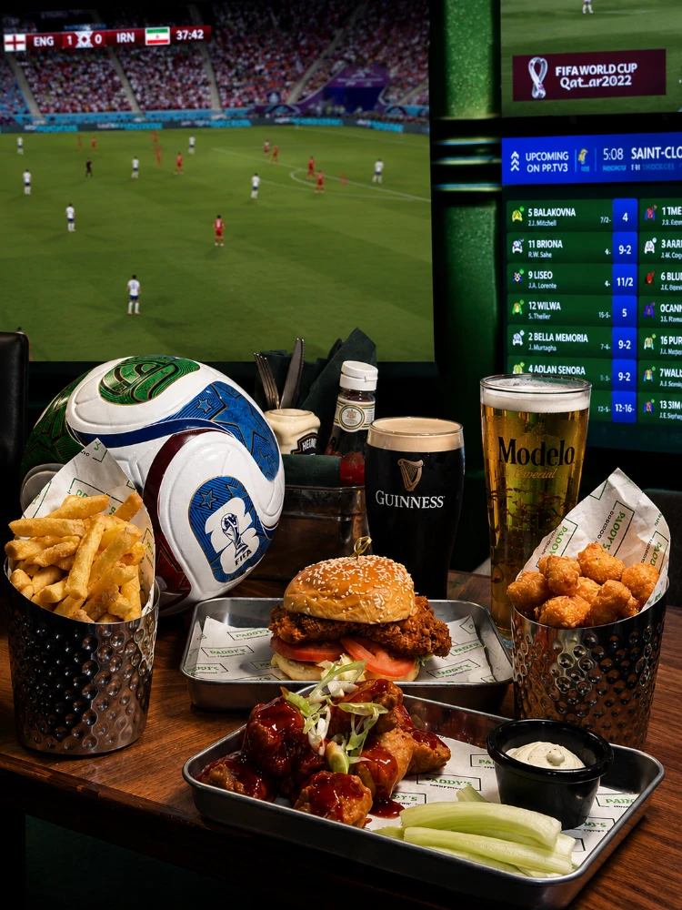 A World Cup football match on TV. In front, a table with fries, a burger, chicken wings, celery, and beers; evoking a lively sports bar vibe.