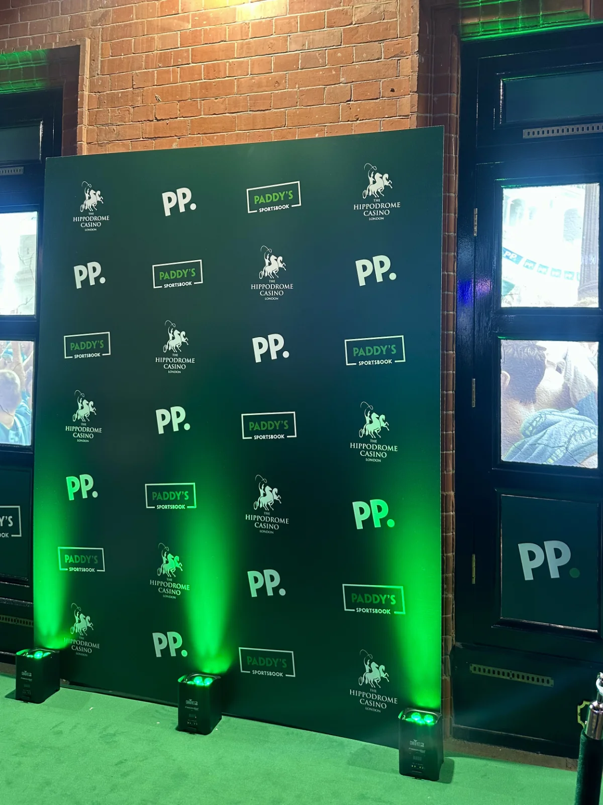 Green-lit event photo backdrop covered with P.P., Paddy’s, and Superdome logos, set against exposed brick beside dark windows and doors.