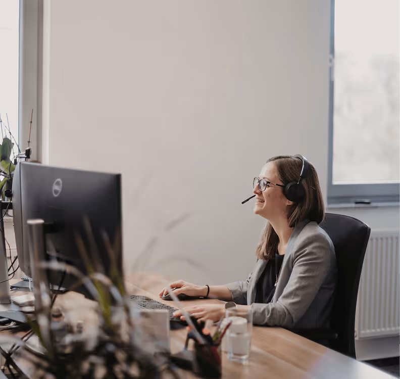 Frau mit Headset sitzt am Schreibtisch, arbeitet am Computer, Büropflanzen im Vordergrund.