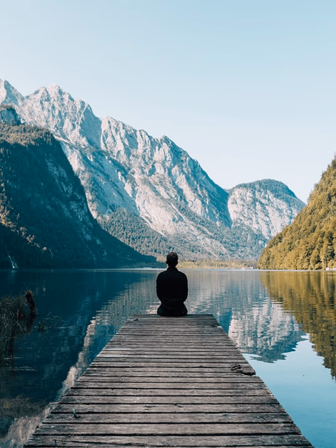 Person sitzt allein auf Steg, blickt auf Bergsee und Alpenlandschaft, klare Spiegelung im Wasser.