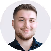 Lächelnder Mann mit kurz geschnittenem Haar und Bart in einem dunklen Hemd vor neutralem Hintergrund.