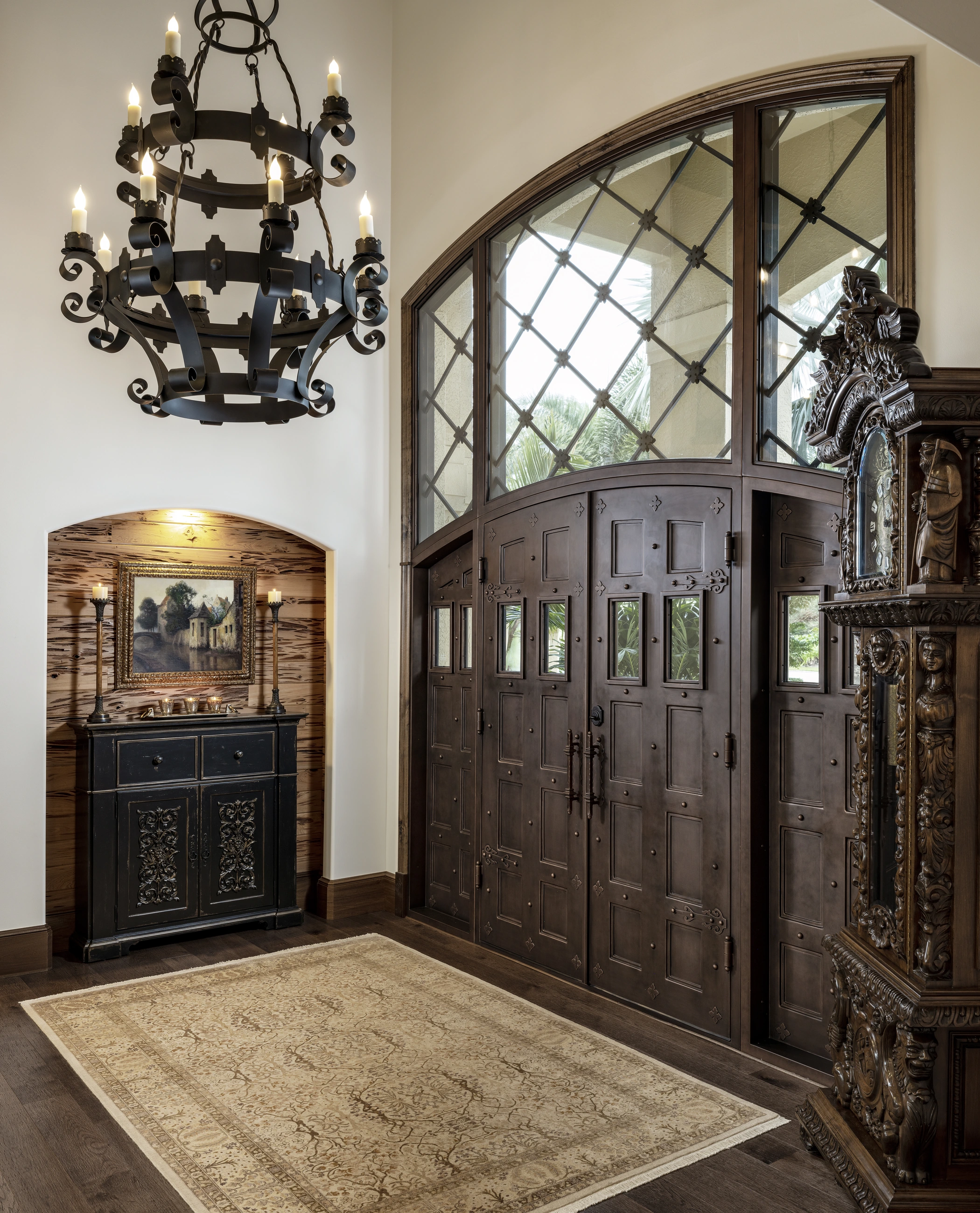 A full view of the entry with a carved cabinet in an illuminated wood-lined arch, a chandelier, tall wooden doors, an ornate clock, and a patterned rug