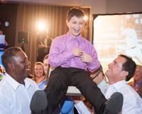 Photo of a Bar Mitzvah boy participating in the traditional Hora where the mean closest to them lift the boy up in the chair while bouncing them up and down.