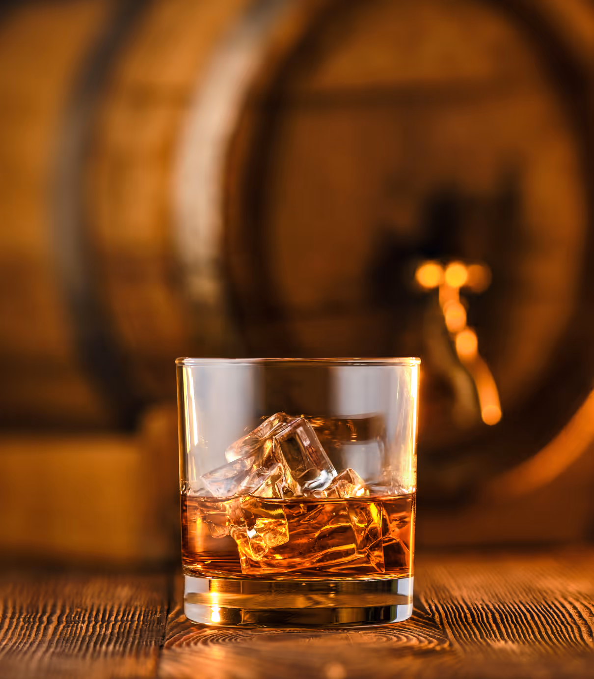 Glass of whiskey with ice cubes on a wooden surface with a blurred barrel background.