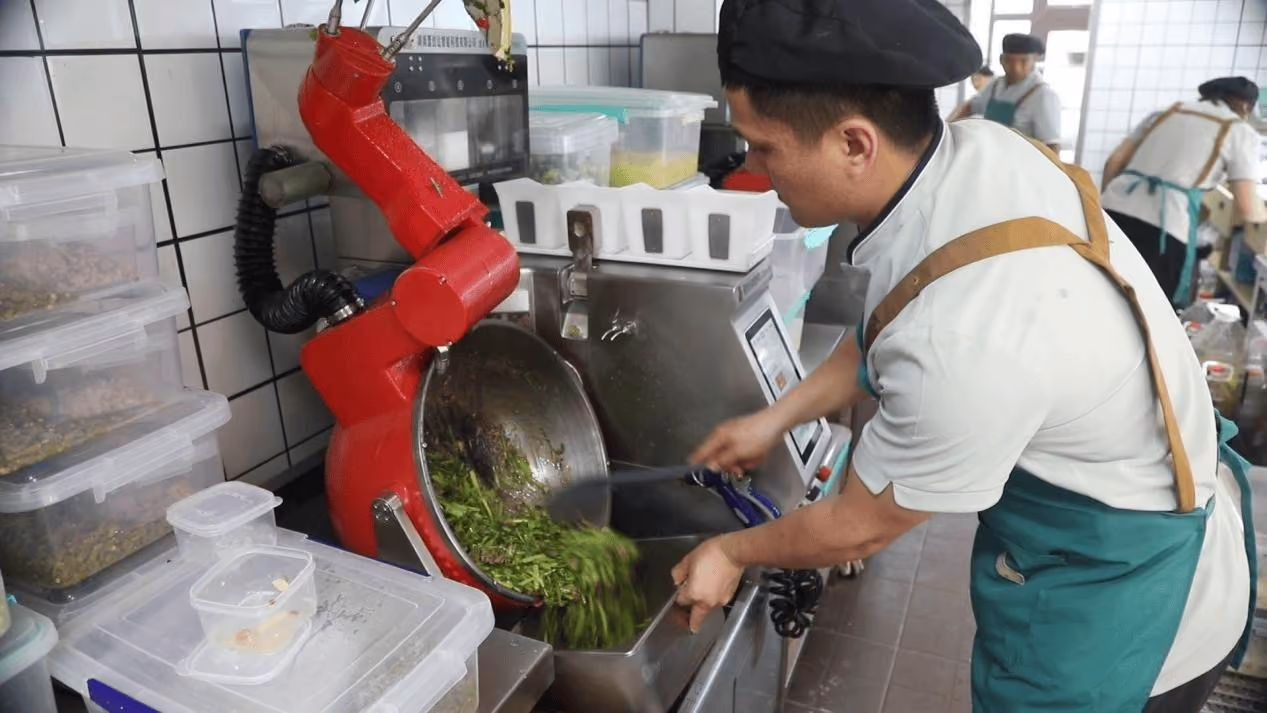 Chef supervising a robotic wok station during ingredient discharge in a commercial kitchen