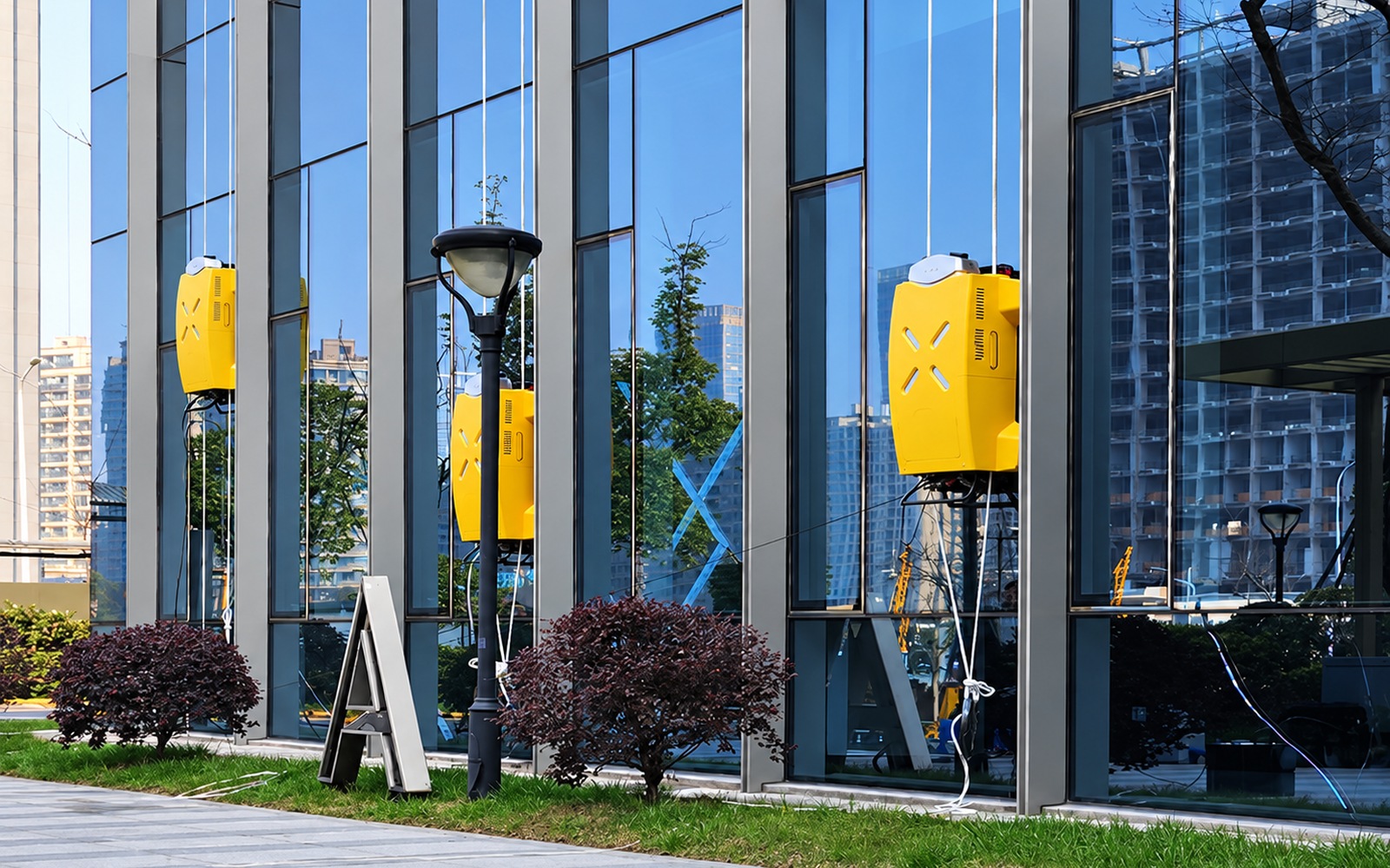 Three unbranded yellow facade cleaning robots in their original operating positions on commercial building glass, with overhead lines and service hoses.