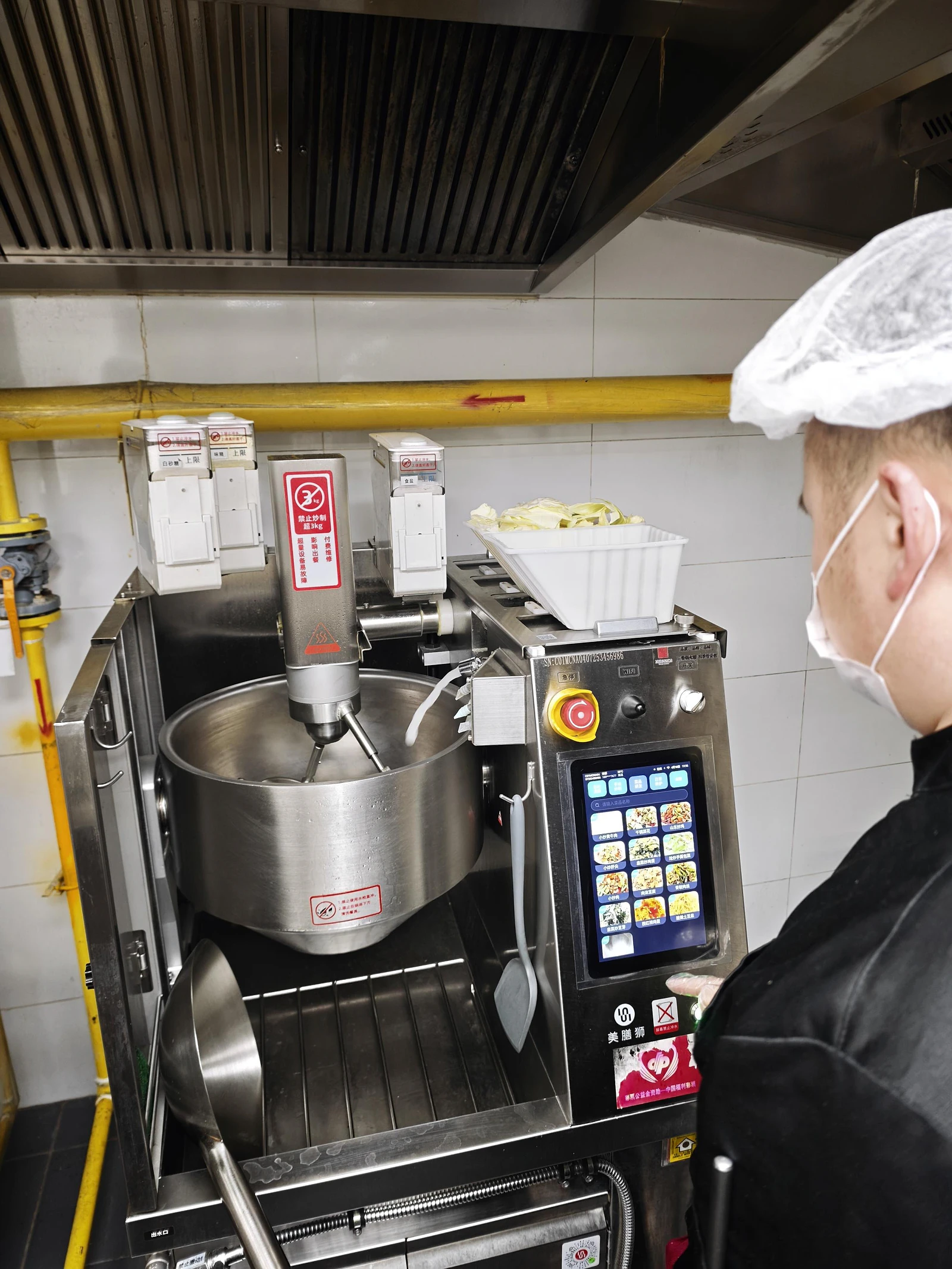 Commercial robotic wok-cooking station operating in the anonymized Singapore kitchen while a staff member monitors the process.