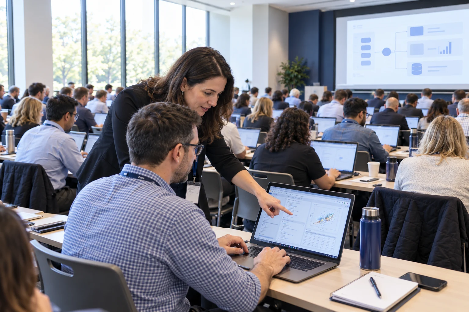 Instructor pointing at an attendee's laptop during a large hands-on workshop, with rows of participants working on computers.