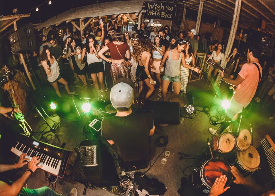 Toma con lente ojo de pez desde el escenario hacia una multitud que baila con energía en un bar rústico al aire libre, con integrantes de la banda visibles en primer plano, incluyendo un tecladista y un baterista.
