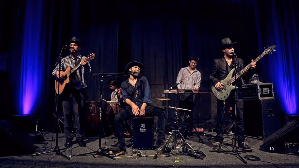 Una banda de folk latino de cinco integrantes toca en un escenario formal con telón azul de fondo — los miembros usan sombreros de ala ancha y tocan guitarra acústica, bajo, cajón, batería y teclado.