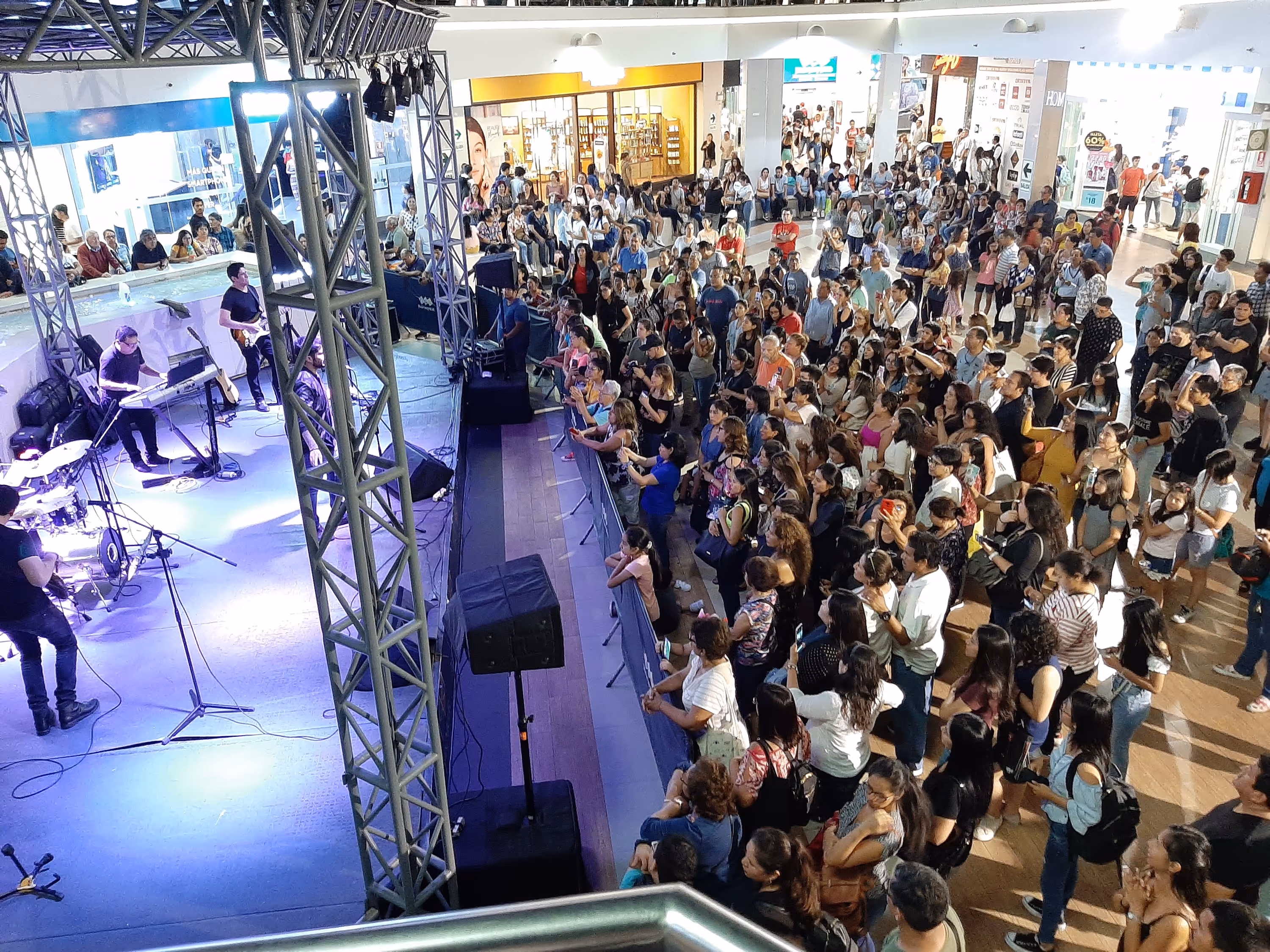 Vista aérea de una gran multitud reunida en el atrio de un centro comercial para ver a una banda en vivo sobre un escenario con torres de metal y luces púrpuras. Muchos asistentes graban con sus teléfonos.